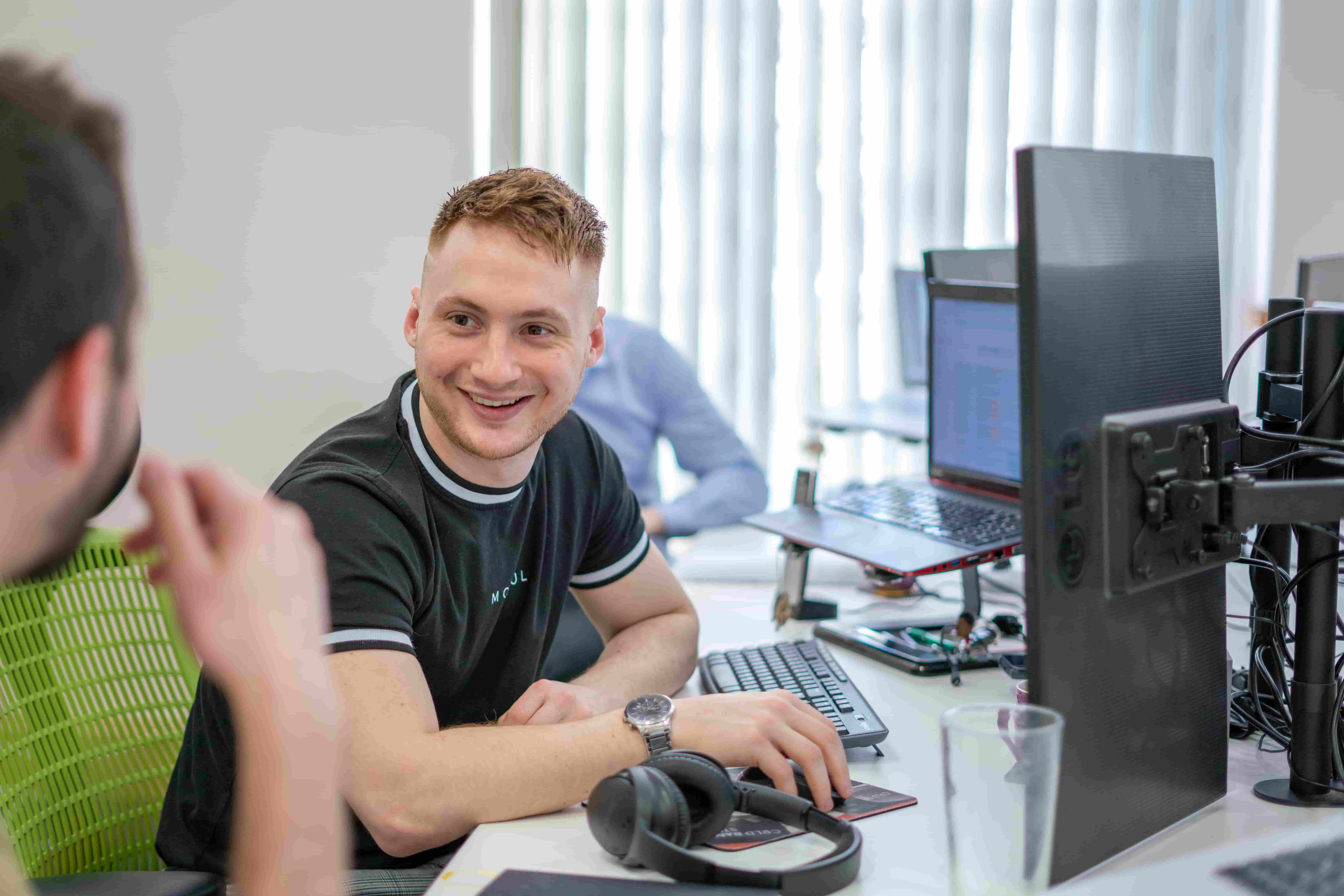 Managing Director Ollie Storey at a computer desk with a laptop and two monitors open, talking to a colleague from left of frame.