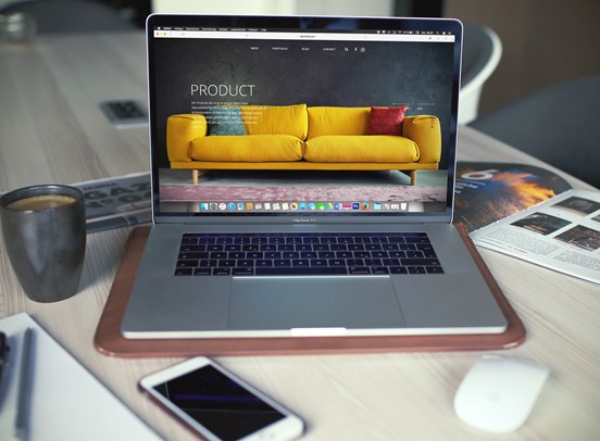 A laptop with a sofa website on it is surrounded by a clutterd table with books, tea, phones and a computer mouse.