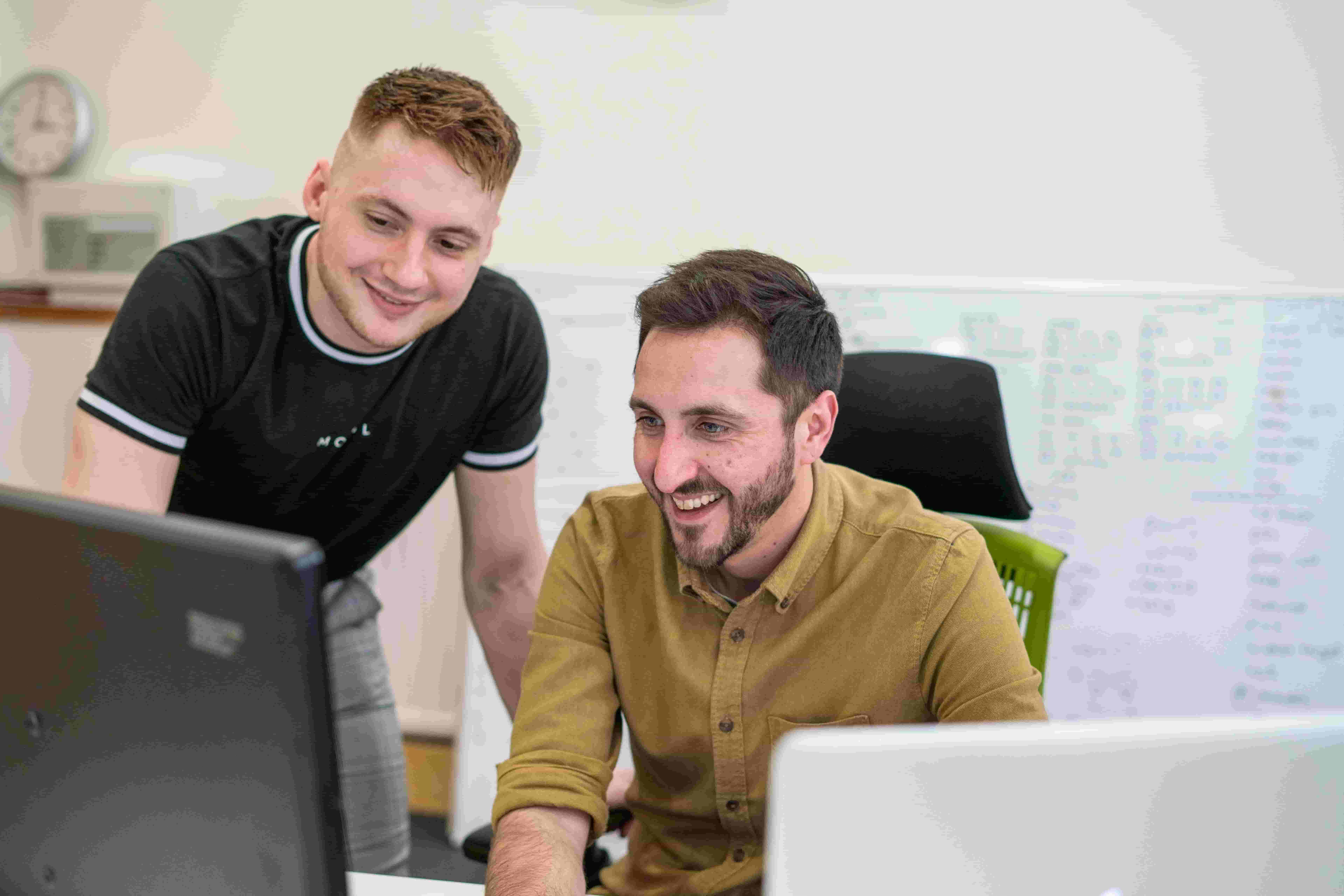 A candid photograph of two Cold Banana employees working on a problem at a computer. In the background is a large whiteboard.