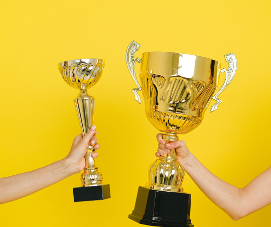 Two trophies being held in frame by two different people against a yellow background