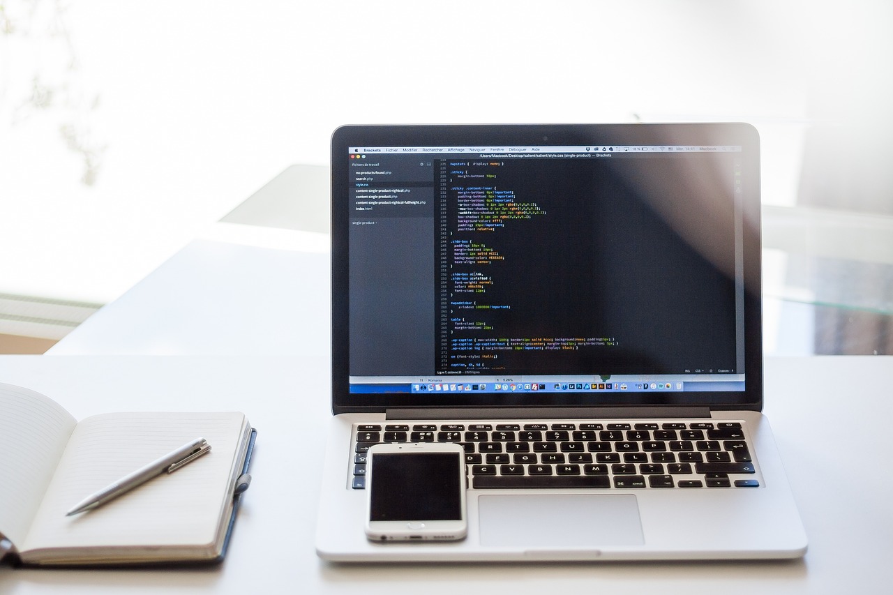 A laptop with a coding window open. A white smartphone is placed upon the keyboard of the laptop. Additionally, a blank notebook lies open to the left of the laptop with a silver pen upon it.