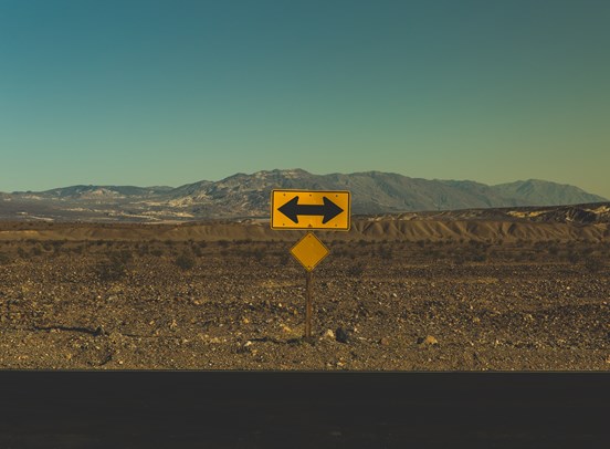A remote mountain landscape in a desert, featuring a yellow sign in the middle with a black two way arrow pointing left and right.