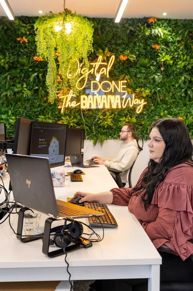 Cold Banana employees working at their desks with a greenery wall in the background featuring a yellow neon sign that reads "Digital Done the Banana Way"