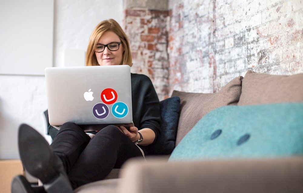 A woman sitting on a sofa with her legs up, reading from a laptop covered in Umbraco stickers.