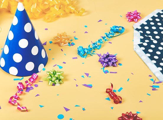 An birthday scene with a blue polka dot party hat, confetti, ribbons and wrapping paper on a yellow background.