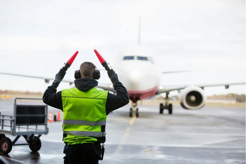 An air traffic controller with marshalling bats directing an airplane on a runway.