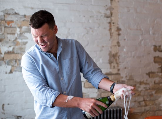 CEO Luke Fribbens opening a bottle of champagne in celebration, with champagne foam flying everywhere.
