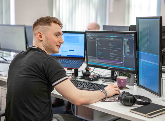 Managing Director Ollie Storey at a computer desk with a laptop and two monitors open.