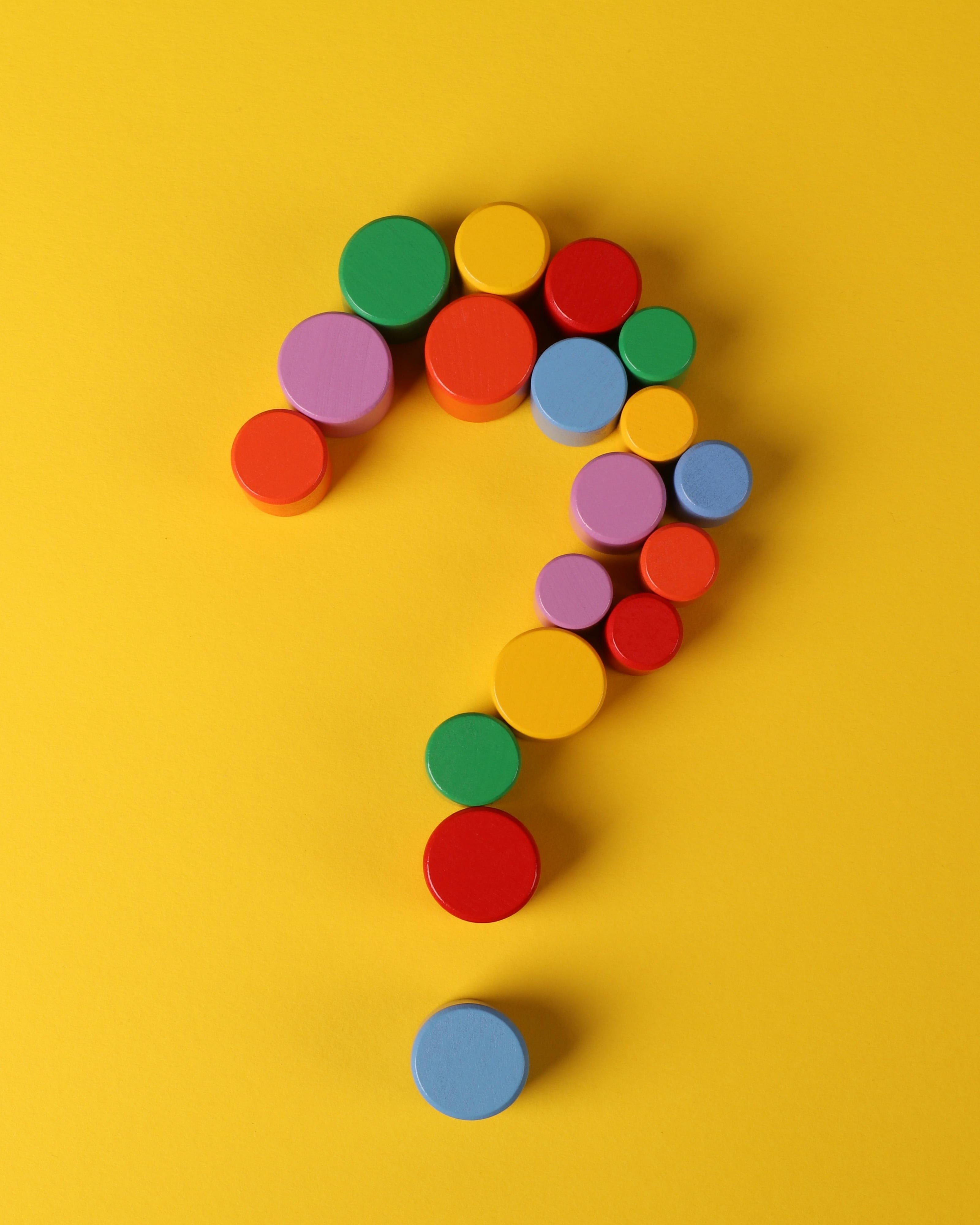 A question mark comprised of multicoloured wooden cylinders, on a yellow background.