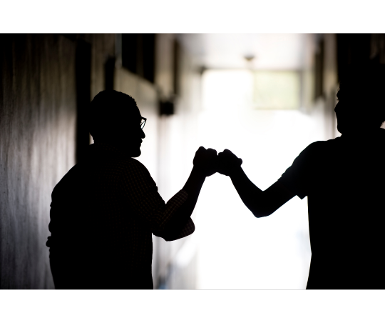 Two people fistbumping one another with the lighting in such a way where they are both silhouetted.