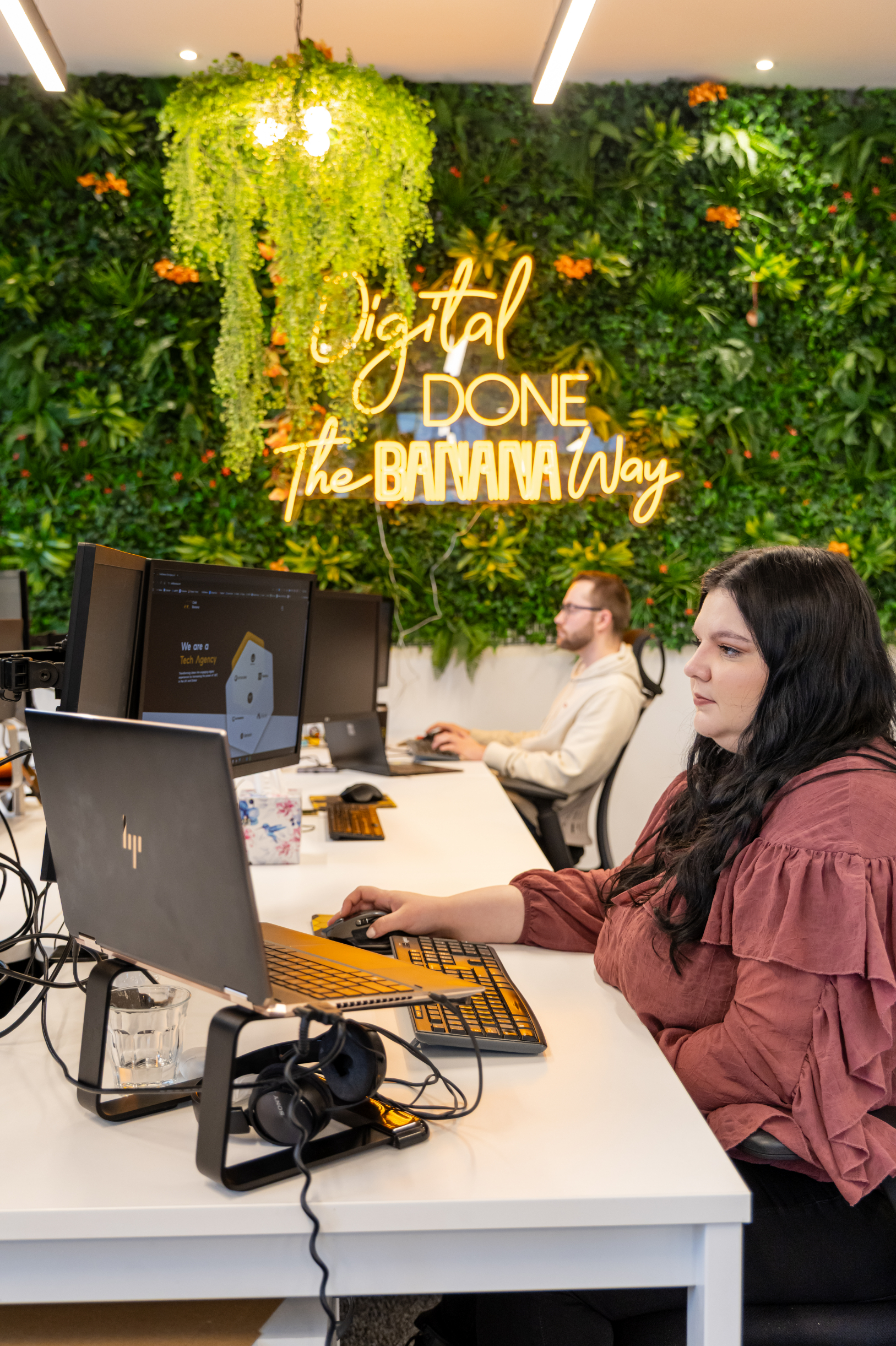 Cold Banana employees working at their desks with the neon yellow sign "Digital Done the Banana Way" in the background on a greenery wall.