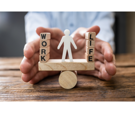 A seesaw made of wooden blocks with the balancing point being a circle. On the blockabove the cylinder is a paper person, with blocks spelling "Work" and "Life" counterweights to one another