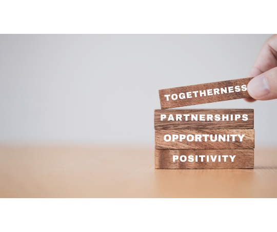 4 blocks stacked on top of one another with a hand placing the fourth block on top. From top to bottom the blocks read as follows "Togetherness, Partnerships, Opportunity, Positivity"