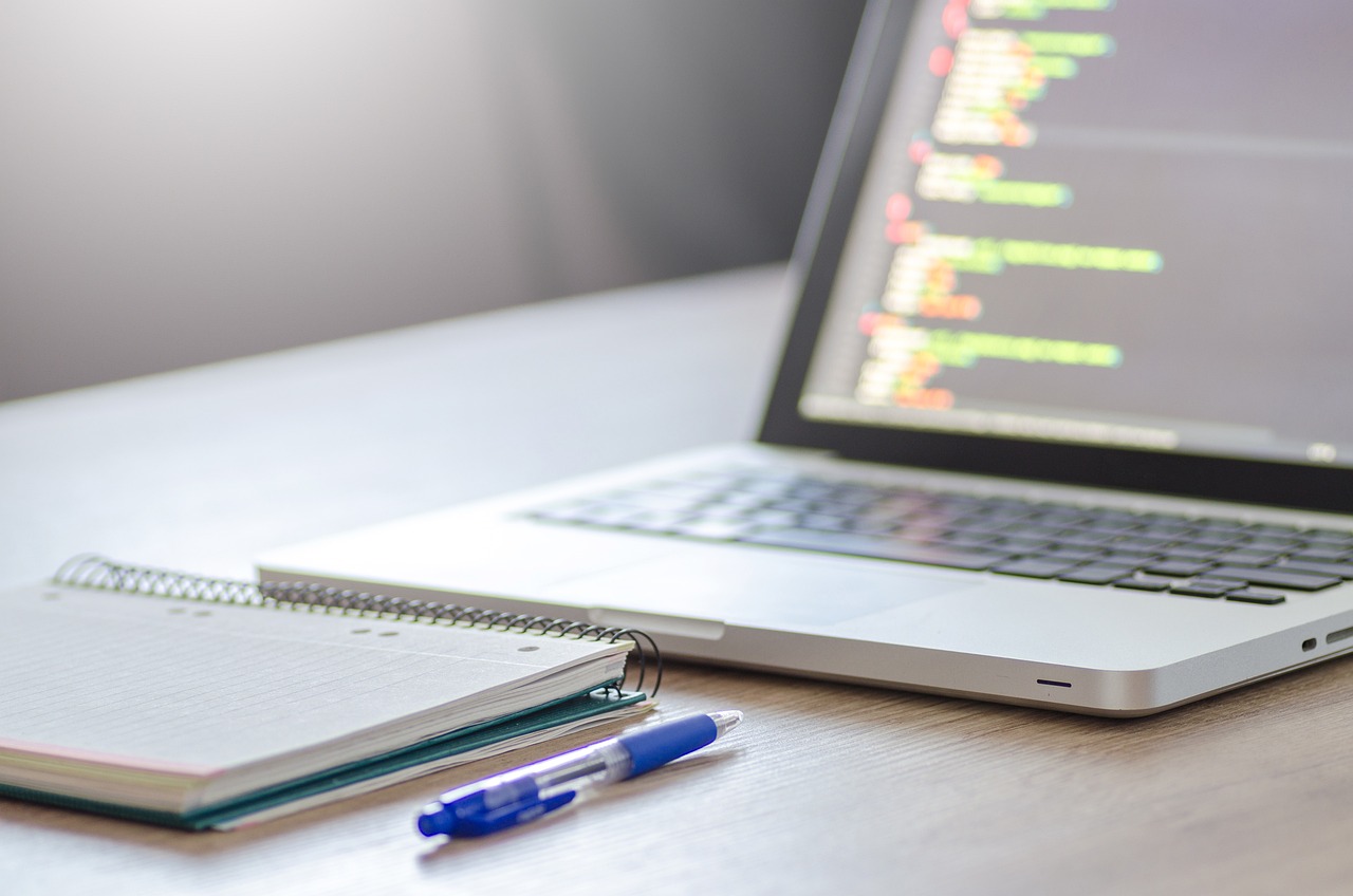 A laptop with a coding window open, with a notebook and blue pen placed before the keyboard.