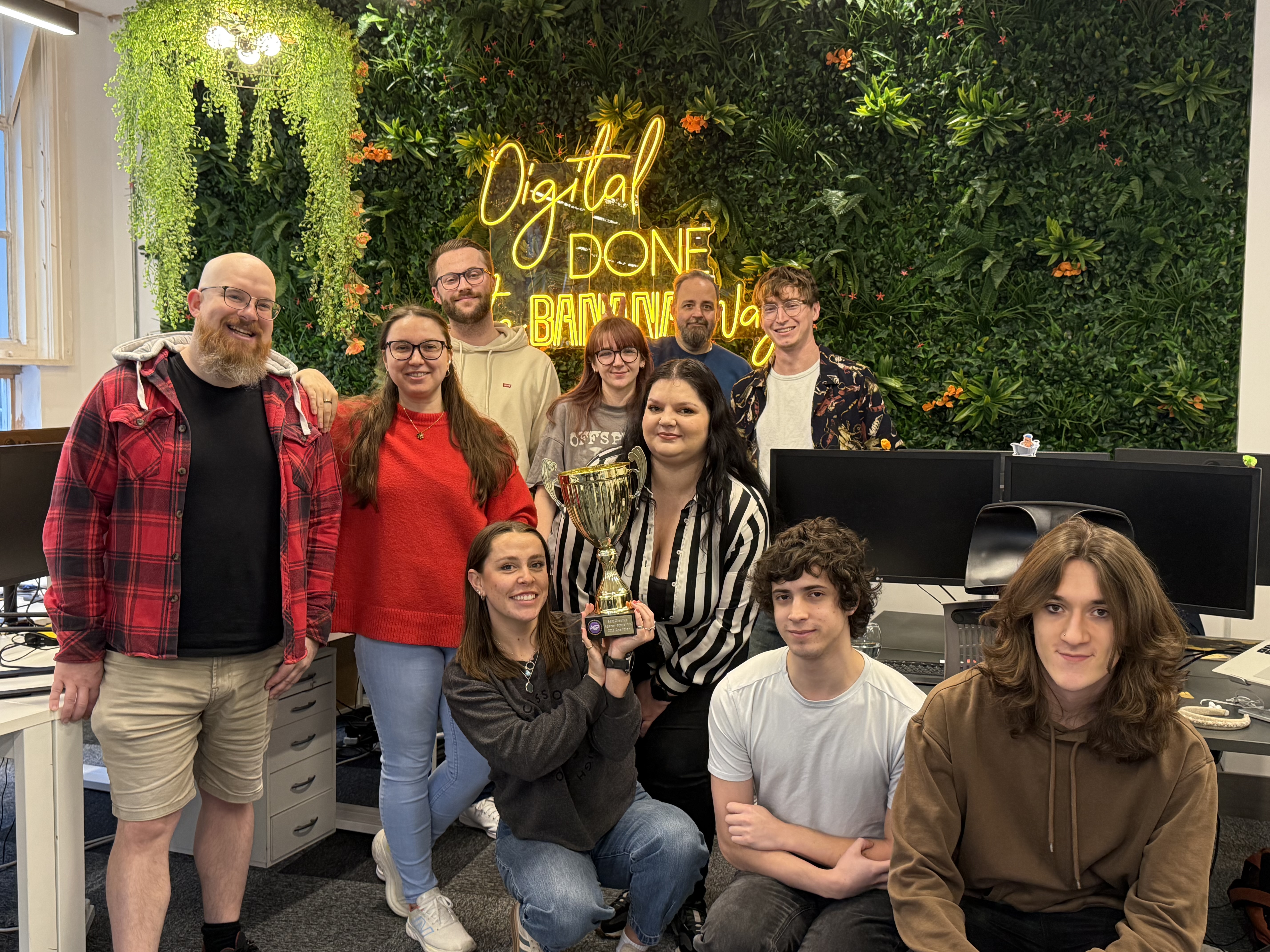 The Cold Banana Bournemouth HQ office team holding the Agency Grand Prix 2025 trophy in front of a wall with greenery and a yellow neon sign reading "Digital Done the Banana Way"