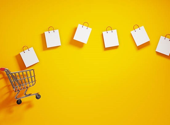 A mini shopping trolley laying on its side on a field of yellow. White shopping bags are floating out of the trolley.